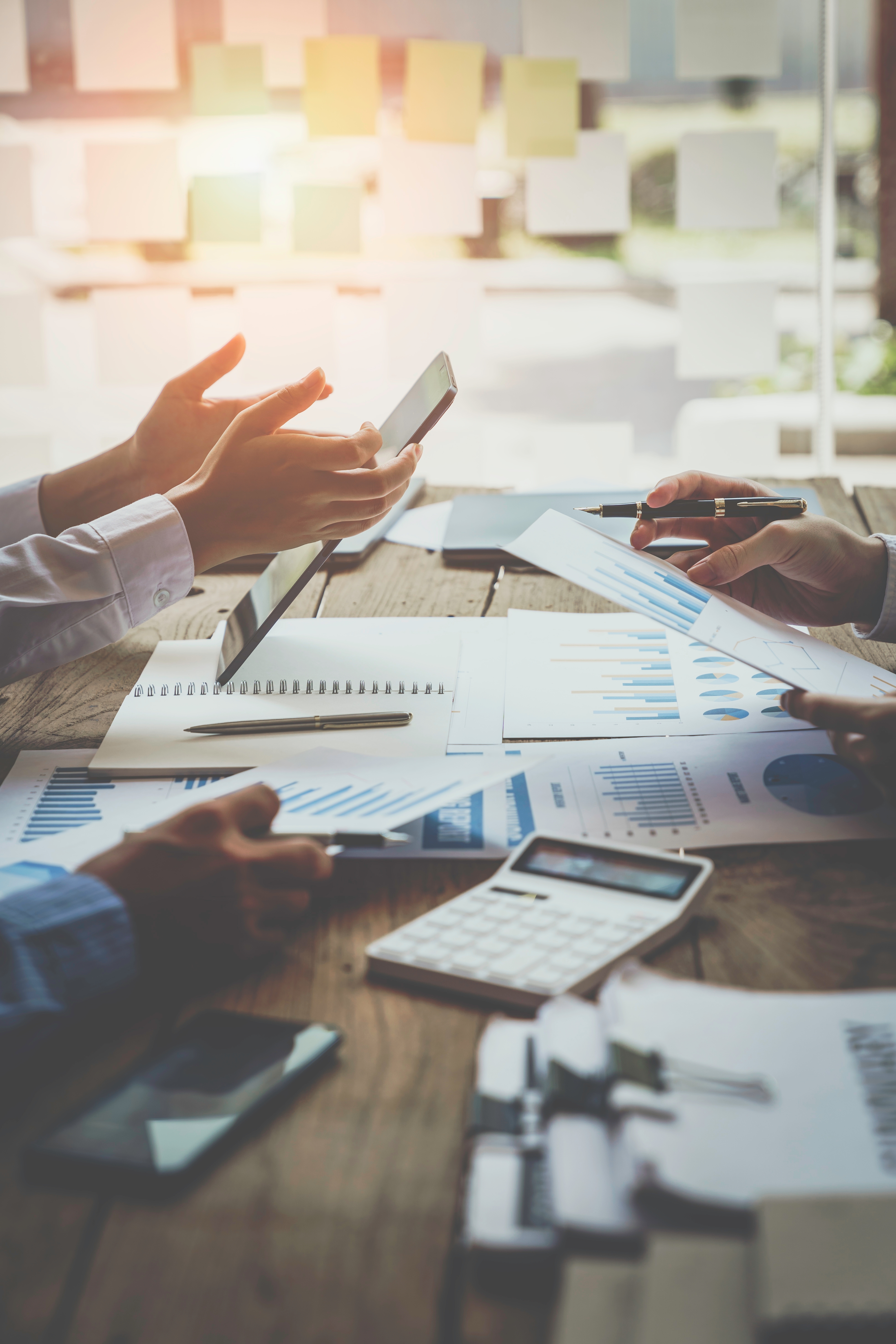A group of hands engaged in a business discussion, holding a tablet and documents with graphs and charts, surrounded by a calculator and stationery on a wooden table.