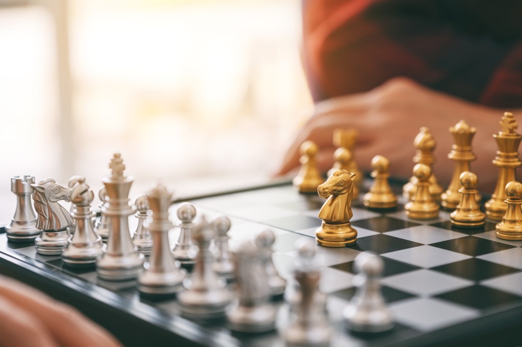 Close-up of a chessboard featuring silver and gold chess pieces, with a player in the background contemplating their next move.