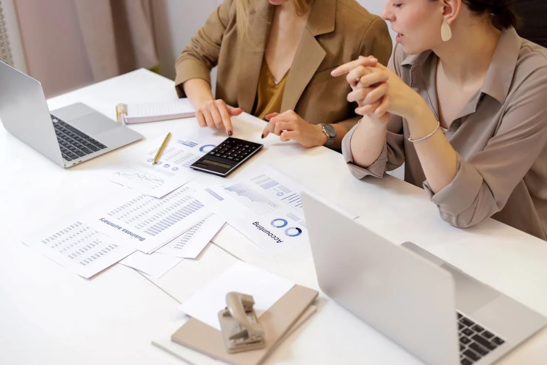 Two women discussing financial documents at a desk, with laptops and a calculator present.